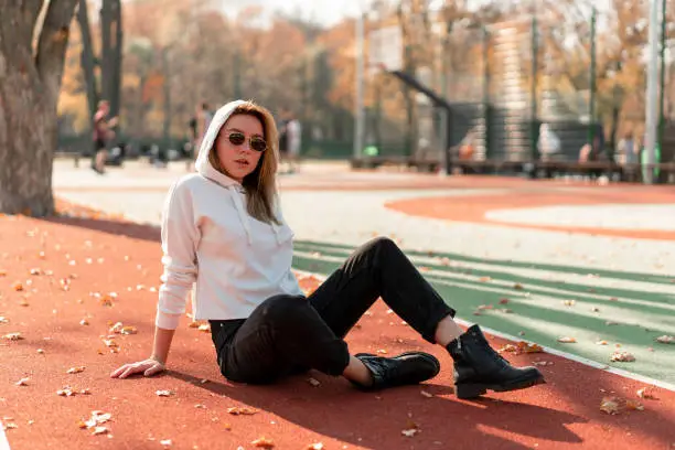Outdoor portrait of young beautiful woman with long in sunglasses and a white hooded sweater sitting on the sportsground track.  youth culture summer pastime