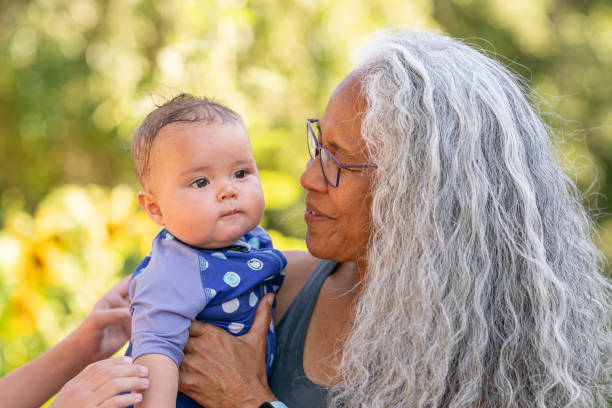 Baby in swimsuit snuggles grandma on a hot summer day A cute baby girl relaxes in her grandmother's arms outside on a warm summer day. The beautiful Pacific Islander senior woman is smiling affectionately at her granddaughter. The baby is dressed in a swim suit and is wet from playing in the kiddie pool in the backyard. legacy concept stock pictures, royalty-free photos & images