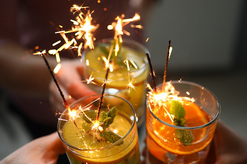 Close-up of friends hands holding three bright delicious cocktails with sparklers. Fruit alcoholic or non-alcoholic beverages. New year party and holiday concept