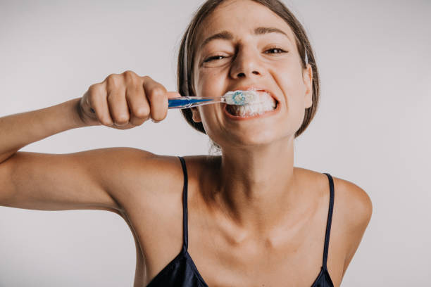 Brushing teeth looks fun A funny photo of a cute young adult brushing her teeth wearing a nightgown, facing the camera. Isolated in a studio with a white background. toothbrush photos stock pictures, royalty-free photos & images
