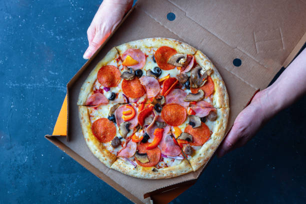 istock Takeaway delivery service. Female hand holds open cardboard box with pizza. Fresh prepared pizza with mushrooms, ham and cheese on a dark background. Fast food concept. Copy space, top view 1272917067