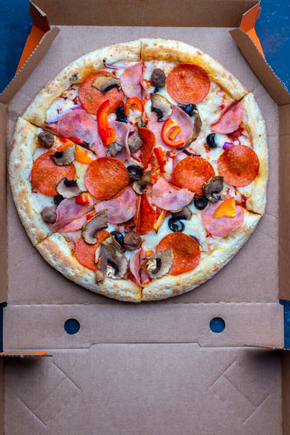 istock Takeaway delivery service. Opened cardboard box with fresh prepared pizza with mushrooms, ham and cheese on a dark background. Fast food concept. Copy space, top view 1272917068