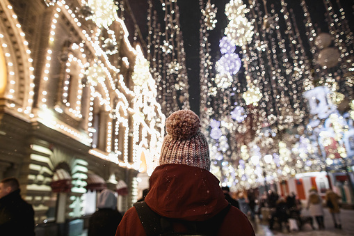 Outdoor night portrait of young fashionable woman in the winter hat with pompom rear view. Magic snowfall effect. Night street illumination in Moscow
