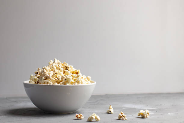 ボウルの横にポップコーンがほとんどない灰色のテーブルの上の白いボウルのポップコーン。 - popcorn snack bowl corn ストックフォトと画像