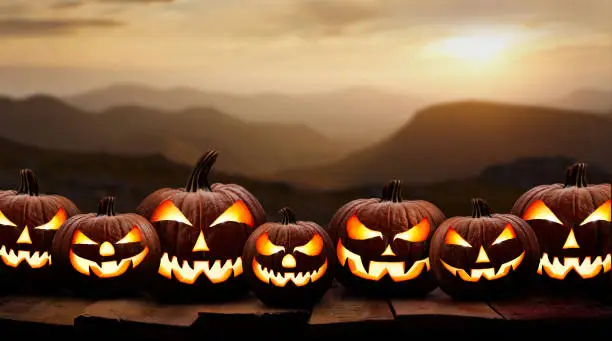 Seven spooky halloween pumpkin, Jack O Lantern, with an evil face and eyes on a wooden bench, table with a sunset mountain background.