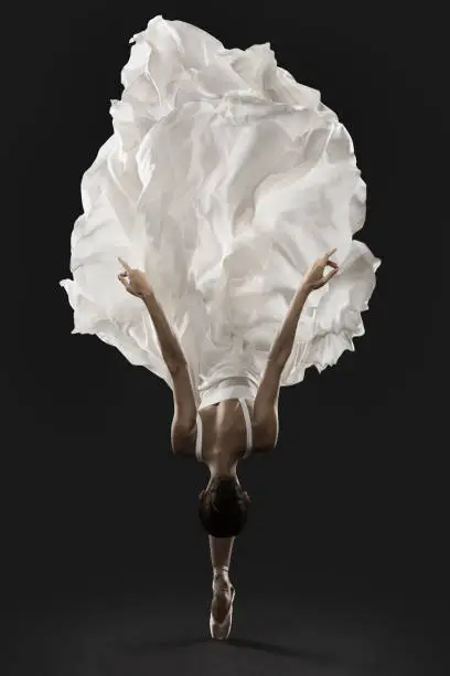 Photo of Ballerina Graceful Jump in White Silk Dress, Ballet Dancer Pointe Shoes in Fluttering Cloth, Black Background