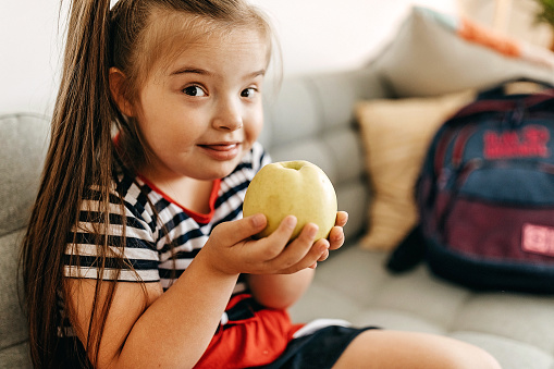 Girl with Down’s syndrome sitting on sofa and eating apple, preparing for school