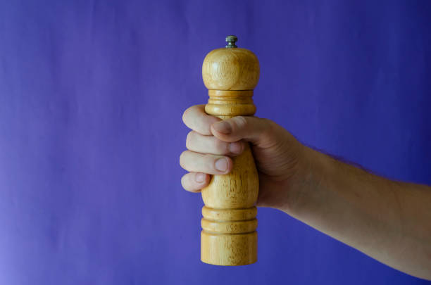 A male hand holds a pepper mill on a blue background. Wooden kitchen fixture for spices. Selective focus.