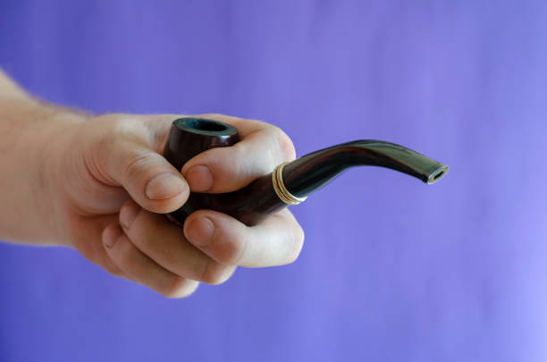 Smoking pipe in hand against the background of a blue wall. An adult male's right hand holds a smoking pipe made from heather or briar root. Monochrome photo. Selective focus.