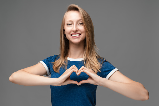 Young Blonde woman, dressed in blue t shirt, making heart symbol by hands on isolated gray background. Human emotions concept.