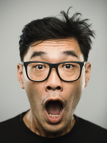 Close up portrait of asian mid adult man with surprised expression against white gray background. Vertical shot of chinese real people with mouth open in studio with black hair and glasses. Photography from a DSLR camera. Sharp focus on eyes.