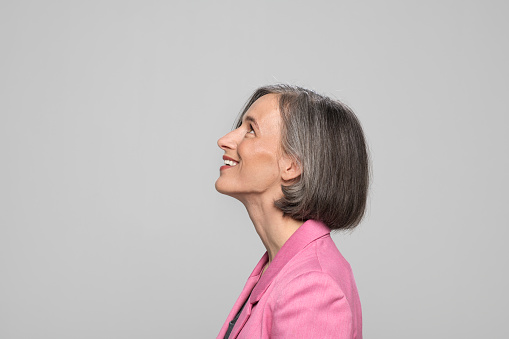 Profile of smiling mature woman looking up while standing against grey background.