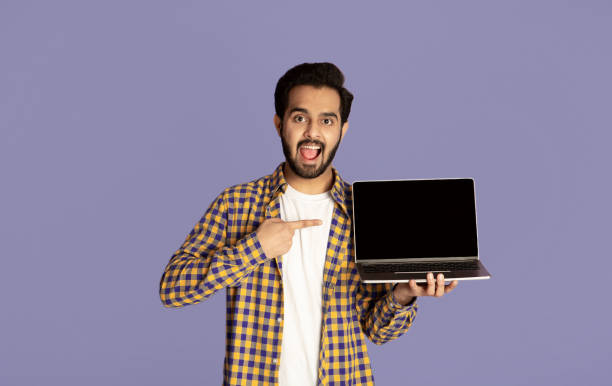 istock Excited Indian man pointing at empty laptop screen over violet background, mockup for website design 1272958588