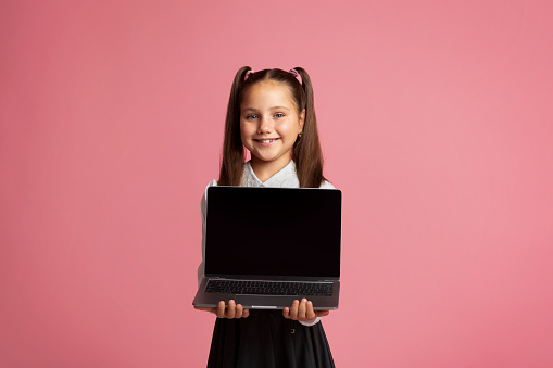 Remote education during quarantine. Smiling girl in school uniform holding laptop with blank screen, isolated on pink background