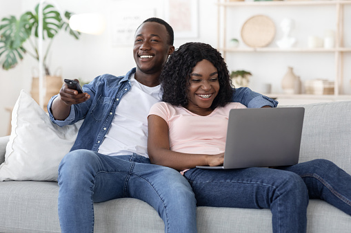 Young black couple watching TV and using laptop, weekend at home