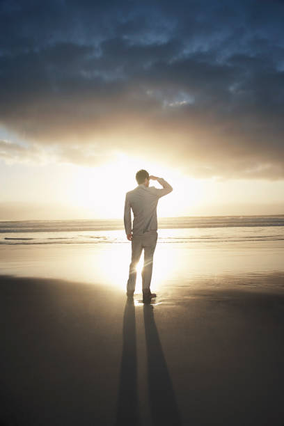 vista traseira do empresário parado na praia olhando para o horizonte ao pôr do sol - looking at view searching looking sea - fotografias e filmes do acervo