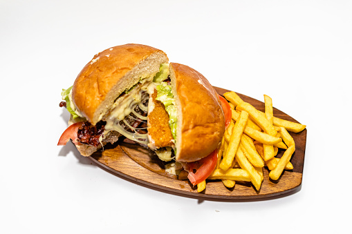 Juicy hamburger with a fried cheese, bacon, tomato and lettuce leaf on a wooden board with french fries to side isolated on white background