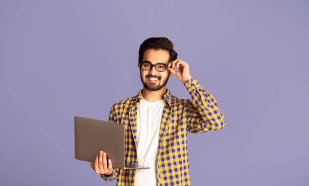 istock Happy Indian guy in glasses using laptop computer for distance work or education on lilac background 1272971719