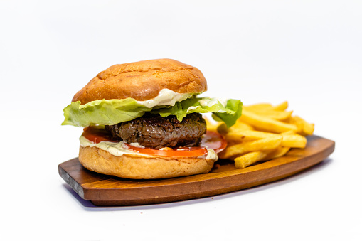 Juicy beef hamburger with a bacon, tomato and lettuce leaf on a wooden board with French fries to side isolated on white background