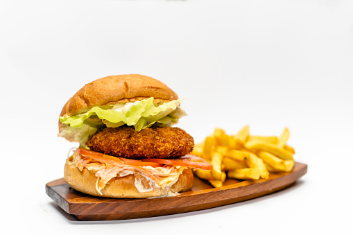 Juicy chicken hamburger with a bacon, tomato and lettuce leaf on a wooden board with french fries to side isolated on white background