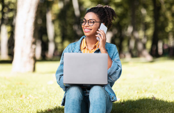 istock Smiling Black Student Girl Speaking On Cellphone Using Laptop Outdoors 1272973018