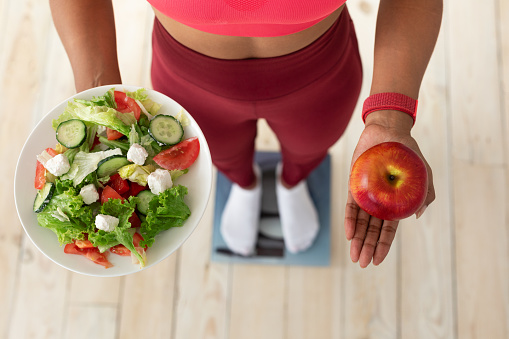 Healthy Diet For Weight Loss. Fit African Woman Standing On Scales Holding Salad And Apple Indoors. Cropped, High-Angle Shot