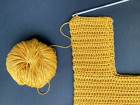 Crocheting. The product is in the process of creation, a ball of mustard-colored yarn and a hook on a black background. Photo from above. Crocheting and knitting, needlework. The concept of creating