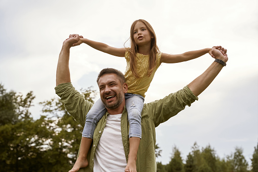 Enjoying time together. Little excited girl having fun with father in park, sitting on his shoulders and smiling. Happy family, parenthood and childhood, Father Day concept