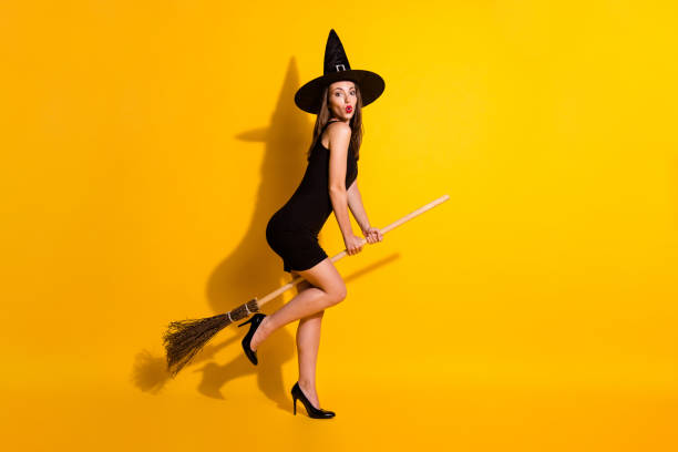 Waist up front view portrait of a mid adult woman dressed up in a witch costume for Halloween, she is smiling and looking at the camera with her hands raised to look scary.