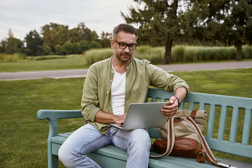 Young handsome man sitting on the wooden bench in park and using laptop, working outdoors. People and nature, freelance and distance work concept