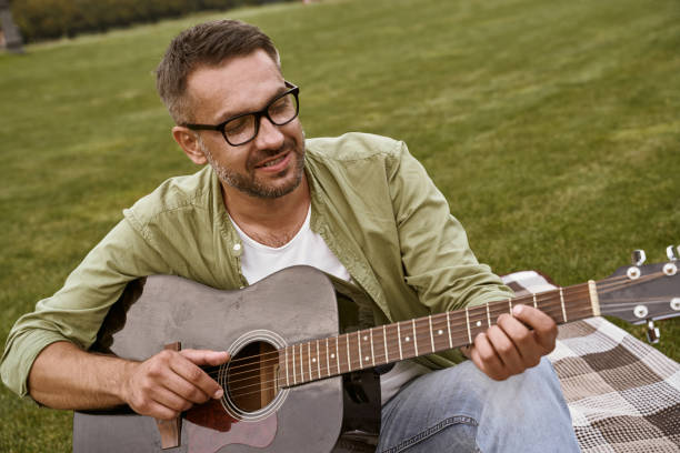istock Young bearded man wearing eyeglasses playing acoustic guitar while sitting on a green grass in park 1272978419