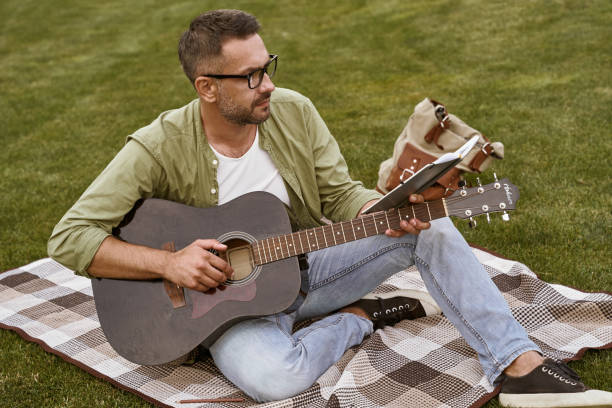 istock Young focused man, male musician wearing eyeglasses reading notes and practicing acoustic guitar while sitting on a green grass in park outdoors 1272978483