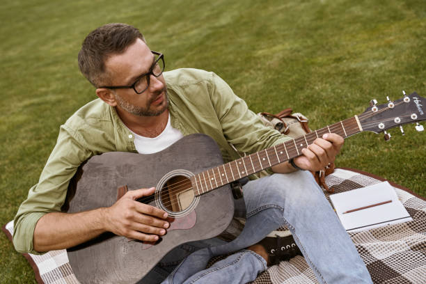 istock Composing music. Young focused man wearing eyeglasses playing acoustic guitar while sitting on a green grass outdoors, male musician working in park 1272978500