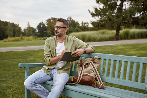 In search of inspiration. Young thoughtful man wearing eyeglasses sitting on the wooden bench in park and making some notes, working outdoors. People and nature, freelance and distance work concept