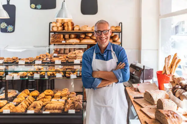 Cheerful mature man with grey hair wearing apron indoor with copy space, standing in bakery while looking at camera with smile.