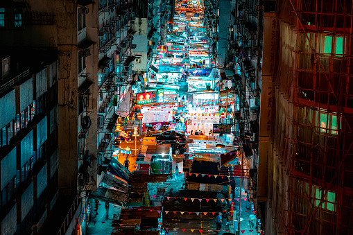 Portrait orientation, overhead view of Hong Kong's Temple Street night-market. The glowing, warm tones of the market street cut through the center of the image, with dark, muted apartment blocks on either side.