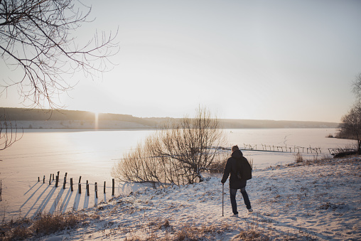 A man with a stick stands looking at a frozen river under ice and snow. Cold harsh winter. All land is covered in white snow. the sun is shining. Disabled man with a backpack.
