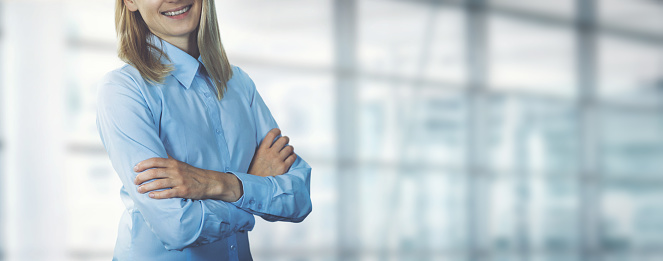 young smiling businesswoman wearing blue blouse standing with arms crossed in modern office. banner copy space