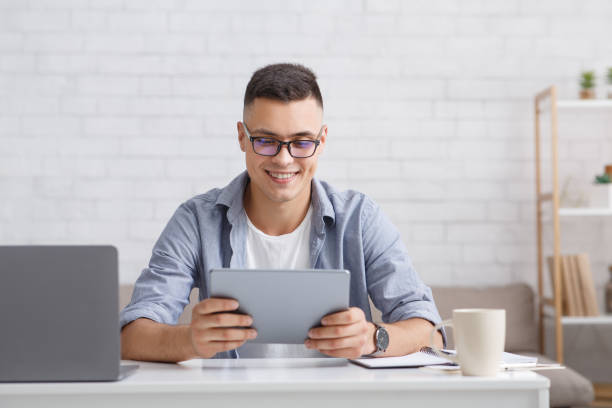 istock Modern hobbies and applications for home. Smiling guy with laptop looks in tablet 1272999592