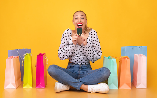 Shopaholic Concept. Portrait of surprised excited woman holding credit card and looking at camera with open mouth, screaming with joy, sitting on the floor at yellow studio background