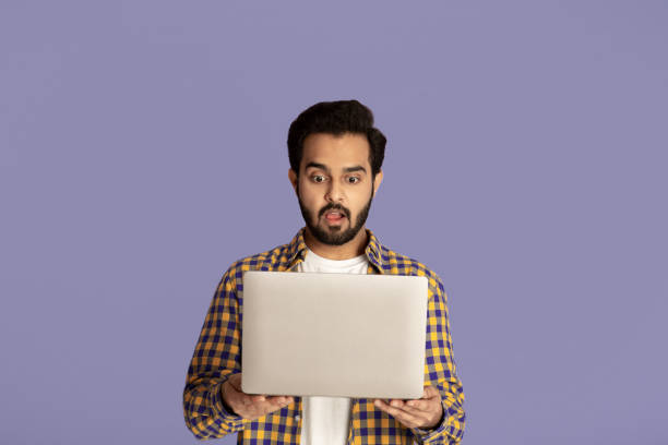 istock Shocked Indian guy staring at laptop screen with open mouth over lilac background 1273001347