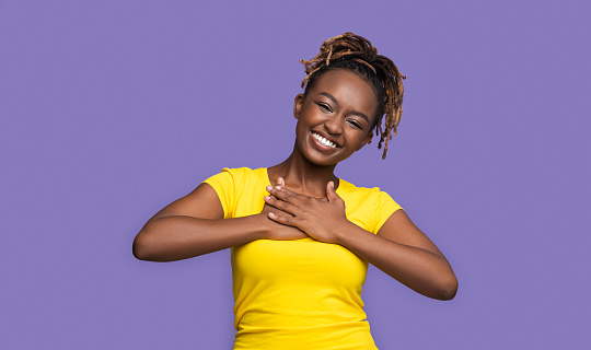 Thankful smiling african girl holding hands on her chest, purple studio background