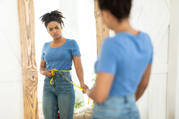 mujer negra infeliz que mide la cintura de pie en el espejo interior - dieting mirror healthy lifestyle women fotografías e imágenes de stock