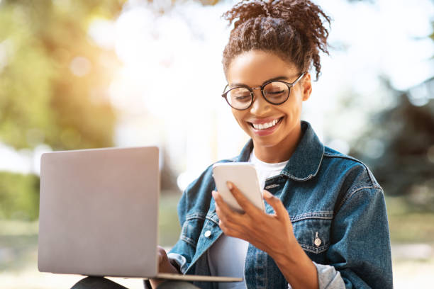 ragazza africana che sms su smartphone imparando usando laptop seduto all'aperto - smiling women glasses assistance foto e immagini stock