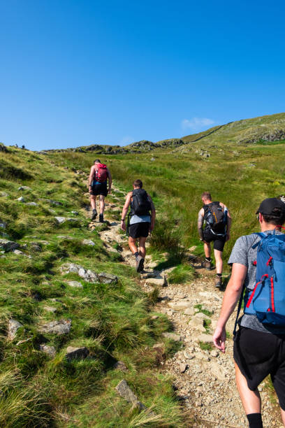 On Our Way To The Top A shot of a group of mid adult men wearing summery hiking clothing and footwear walking up a path. They are in a rural, mountainous setting on a summers day. striding edge stock pictures, royalty-free photos & images