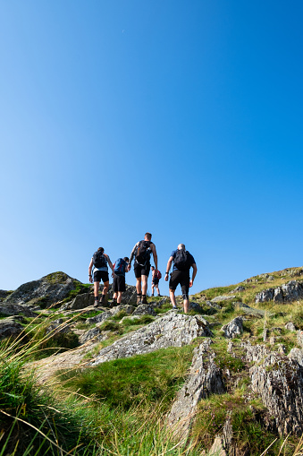 A shot of a group of mid adult men wearing summery hiking clothing and footwear walking up a path. They are in a rural, mountainous setting on a summers day.