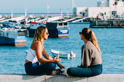 Cascais, Portugal - September 16, 2020: Two women chat while sitting across from Cascais Bay with traditional wooden fishing boats on a sunny day