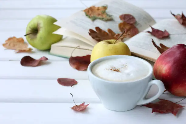 white cup with frothy cappuccino on the background of apples, a book and fallen leaves