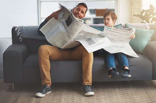 Shot of a carefree little boy and his father reading the newspaper while being seated on the sofa at home during the day