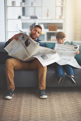 Shot of a carefree little boy and his father reading the newspaper while being seated on the sofa at home during the day